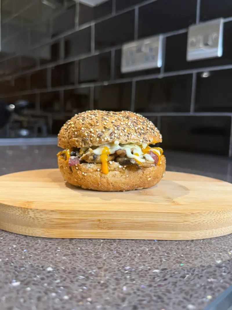 A seeded bagel filled with mushrooms and melted cheese on a wooden board in a tiled kitchen
